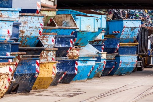 A row of large metal skip containers positioned along a paved area, with some showing signs of rust and wear. The skips are primarily painted in shades of blue, with a few featuring yellow and black accents, and have striped reflective markings on their lower edges for visibility. They are lined up close to each other, with some stacked slightly unevenly, and contain various types of waste materials, including cardboard and mixed debris, visible at the top edges. The environment appears to be an industrial or commercial waste collection site, with a dark vehicle partially visible behind the skips on the right side. The lighting is natural, indicating an outdoor setting, and the overall scene suggests a typical rubbish removal setup in the Chesham area, managed by House Clearance Chesham for waste disposal or clearance services.