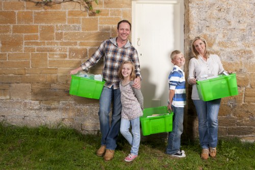 A family of four standing outdoors in front of a stone wall, holding bright green plastic boxes used for rubbish and waste collection. The father, dressed in a checked shirt and jeans, is smiling at the camera while holding a box in his right hand. His daughter, wearing a grey patterned top and jeans, stands close to him and holds onto another green box. To the right, two children—a boy in a striped blue and white shirt and a girl in a white blouse—each hold green boxes, with the boy's hand resting on his box and the girl holding hers by her side. The woman, dressed in a white top and jeans, stands on the far right, also holding a green recycling or rubbish container. They are positioned on a grassy surface with the stone wall behind them, which is part of a residential property, suggesting they are preparing for or have completed rubbish removal. The scene is well-lit with natural daylight, reflecting a typical domestic rubbish collection setting. The image supports services related to household waste or house clearance in Chesham, close to the inferred postcode area, illustrating an organized approach to rubbish disposal for families in the local area.