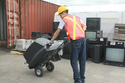 A person wearing a yellow safety helmet, a high-visibility vest with orange and yellow reflective stripes, a white t-shirt, and blue jeans is pushing a black wheeled waste container or bin on an outdoor paved surface. The individual is using both hands to grip the handle of the bin, which appears to be made of durable plastic material. Behind them, there are several old electronic appliances, including televisions, radios, and other electronic equipment, arranged in a semi-organized manner against a backdrop of a rust-colored shipping container and a makeshift shelter made of white tarpaulin. The setting suggests a waste collection or rubbish removal operation, possibly in a storage yard or disposal area for electronic waste. The natural daylight indicates a clear or overcast day. This scene is typical of the rubbish removal services provided by House Clearance Chesham, serving areas near Chesham and the surrounding postcode region.