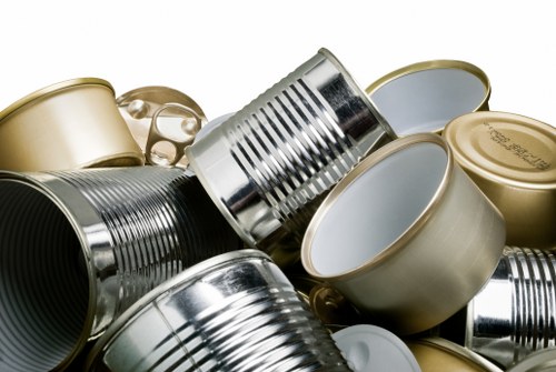A close-up image showing a collection of used metal cans, including both aluminium and steel varieties, piled together on a flat surface. The cans are of various sizes and shapes, with some opened and others with intact, smooth, and reflective surfaces. The metallic textures include ribbed sides on some cans and smooth, shiny finishes on others, with colours primarily in silver, gold, and muted tones. The background is not visible, focusing entirely on the canned objects, which are arranged in a seemingly haphazard manner, indicative of waste collection for rubbish removal services. The scene highlights typical household or commercial waste, reflecting the type of items handled by a company like House Clearance Chesham in the local area, linking visually to their rubbish clearance offerings in Chesham and nearby regions. The lighting is even, emphasizing the metallic sheen and textures of the cans, creating a neutral and factual depiction of recyclable waste materials waiting to be collected and disposed of.