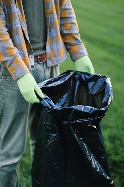A person wearing a white glove is holding a large white polypropylene sack filled with garden debris, including green leafy branches, small twigs, and scattered dry leaves, situated on a concrete or g