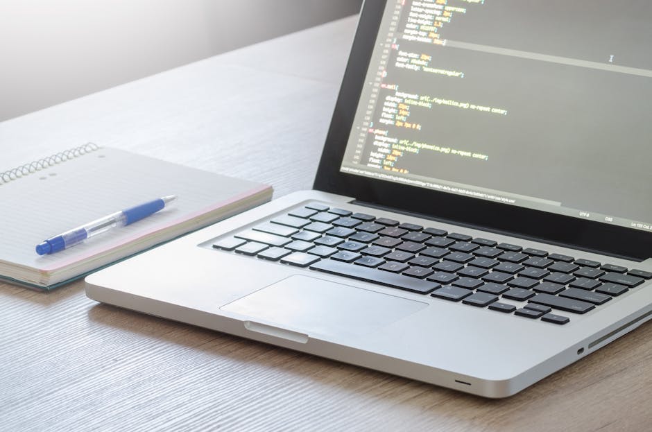 A close-up view of a silver laptop positioned on a light wooden desk, displaying lines of colourful code on a grey background. To the left of the laptop, an open spiral-bound notebook with a white gri