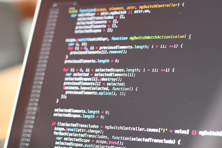 A close-up view of a silver laptop positioned on a light wooden desk, displaying lines of colourful code on a grey background. To the left of the laptop, an open spiral-bound notebook with a white gri